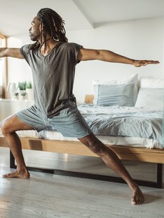 Black man in Warrior II pose in front of his bed at home