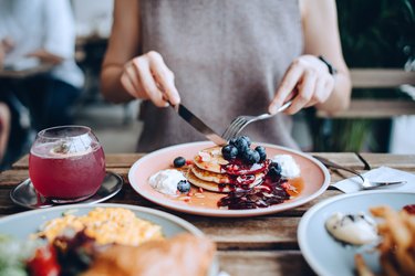 Close view of a person wearing a grey tank top cutting into a stack of pancakes with blueberries, with a brunch cocktail next to their plate
