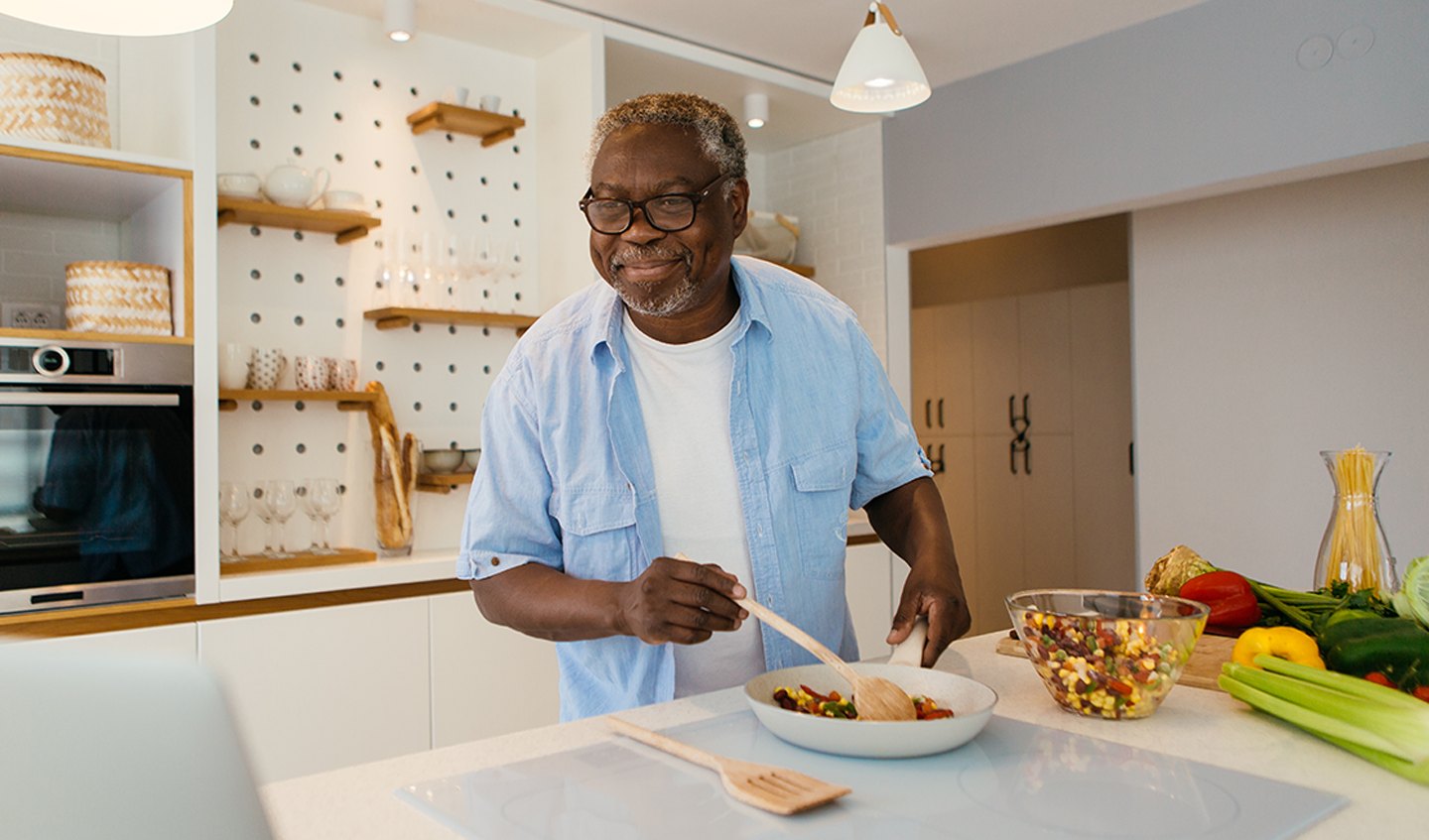 Older man wearing glasses and a light blue shirt mixing together ingredients in his tidy kitchen while watching a cooking video on his laptop