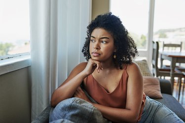 A person with curly hair wearing an orange tank top sitting on their couch looking out the window, depressed in the summer