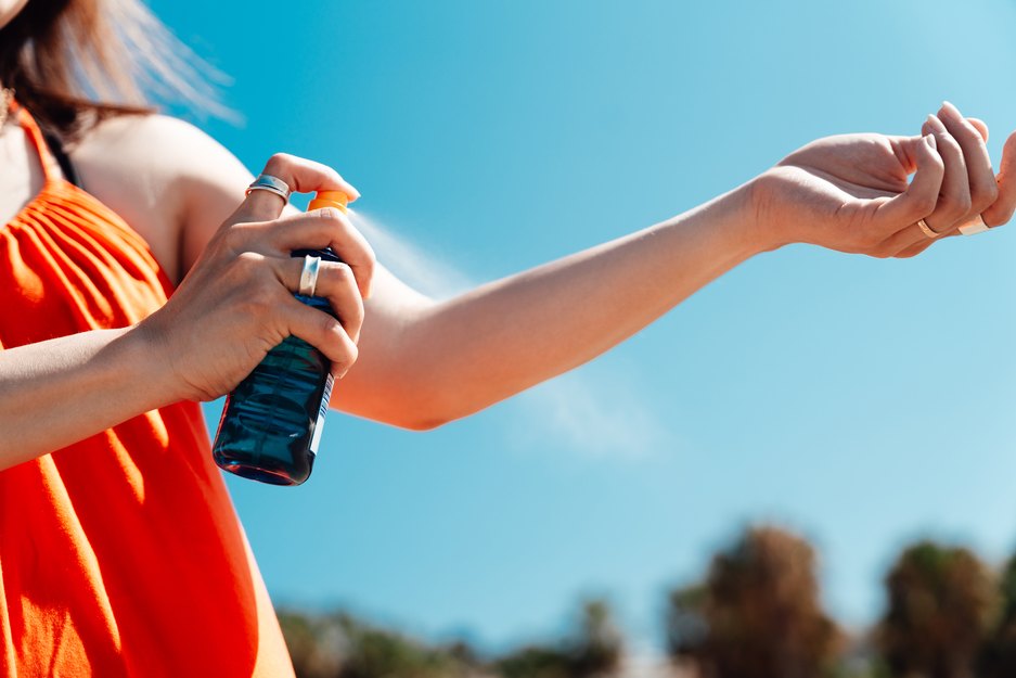 Close-up of a person in an orange tank top spraying sunscreen on their arm on a sunny day
