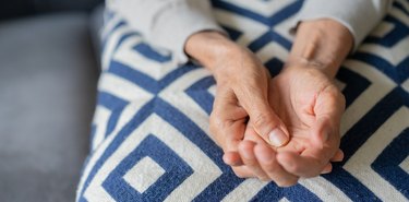 Close up of person sitting on a grey couch wearing a blue and grey criss-cross patterned skirt massaging their hand to relieve pain from arthritis in the fingers