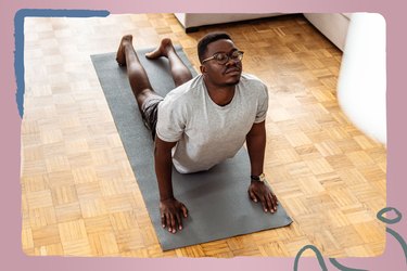 A young adult with short hair wearing glasses and grey workout clothes closes their eyes and holds cobra pose on a dark grey yoga mat at home as part of the 30-Day Yoga Challenge