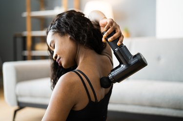 Person using a massage gun on their back and shoulders sitting on the floor at home