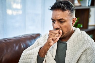 a person with short black hair wearing a blanket and coughing into their hand