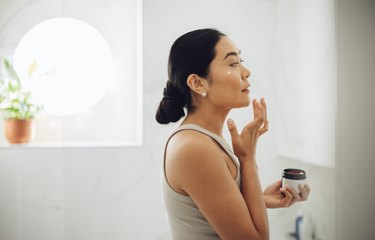 a person wearing a grey tank top with long black hair in a bun applying face cream in the bathroom mirror