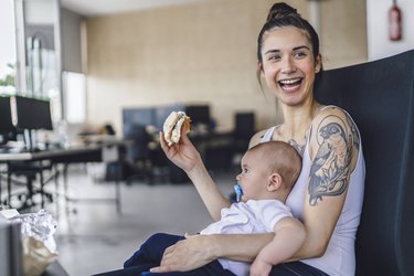 a parent with brown hair in a bun wearing a tank top with arm tattoos sitting in an office with a baby on their lap, eating a sandwich